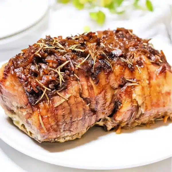 A luscious pork roast garnished with herbs graces a pristine white plate. The meat boasts a glazed, caramelized surface, exuding mouthwatering aromas. In the background, hints of greenery and soft white light create an inviting dining atmosphere.