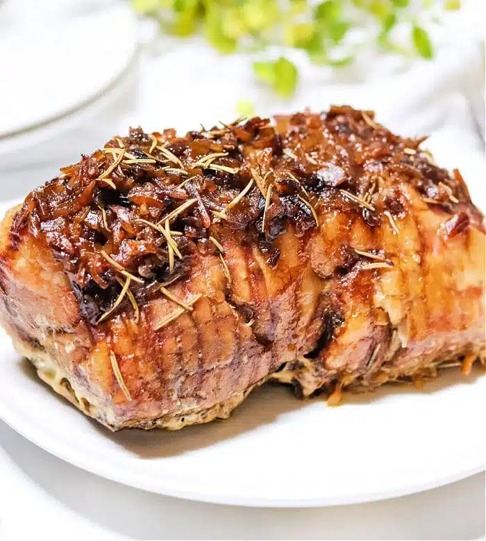 A cooked roast beef garnished with herbs and caramelized onions sits on a white plate, reminiscent of a hearty slow cooker pork roast. The meat appears juicy and is browned on the outside. In the background, there are hints of greenery and a blurred white object.