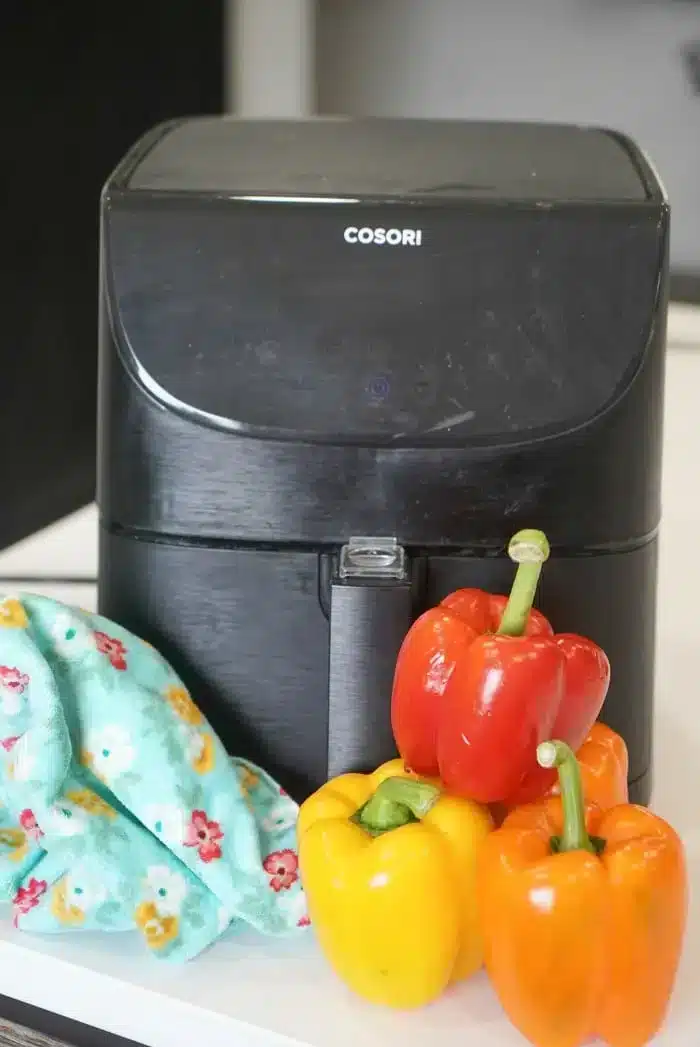 A sleek black air fryer sits proudly on the countertop, flanked by vibrant bell peppers in red, yellow, and orange. Nearby, a blue patterned cloth with a floral design adds a touch of elegance to this colorful culinary scene.