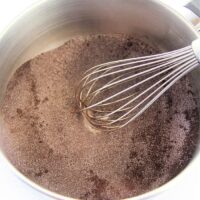 A whisk stirs the homemade brownie mix, blending the dry cocoa powder effortlessly in a metal bowl.