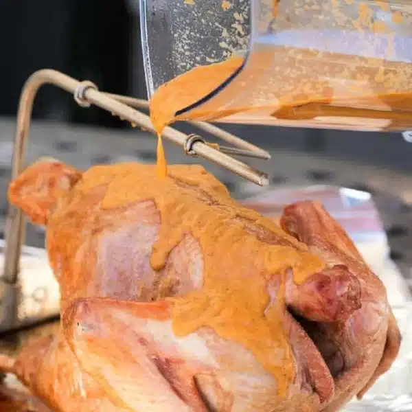 A raw turkey is being coated with a thick, buttery orange marinade poured from a blender. The turkey rests on a metal grilling rack placed on a foil-covered surface, perfect for absorbing the flavorful rub.