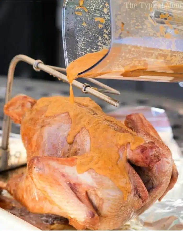 A raw turkey is being coated with a thick, buttery orange marinade poured from a blender. The turkey rests on a metal grilling rack placed on a foil-covered surface, perfect for absorbing the flavorful rub.