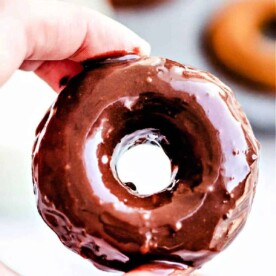 Close-up of a hand holding a glazed donut with blurred baked pumpkin donuts in the background.