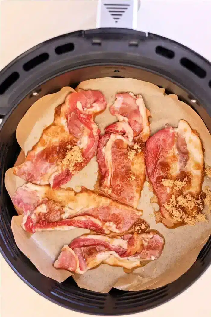 Slices of raw bacon, marbled with fat and kissed with maple syrup, are arranged on parchment paper inside an air fryer basket. The strips appear to be sprinkled with brown sugar, creating the perfect base for candied bacon.