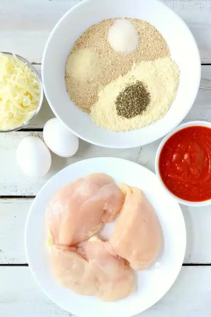 A flat lay of cooking ingredients for air fryer chicken parm: raw chicken breasts on a white plate, two eggs, shredded cheese, a bowl of breadcrumbs mixed with seasonings, and a bowl of red sauce, all placed on a white wooden surface.