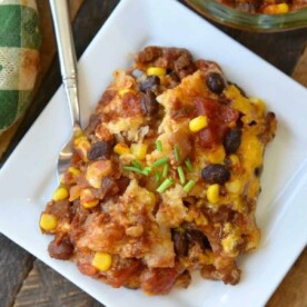 A square plate with a serving of cheesy taco casserole, reminiscent of an enchilada lasagna, topped with chives, sits next to a dish and a green napkin.