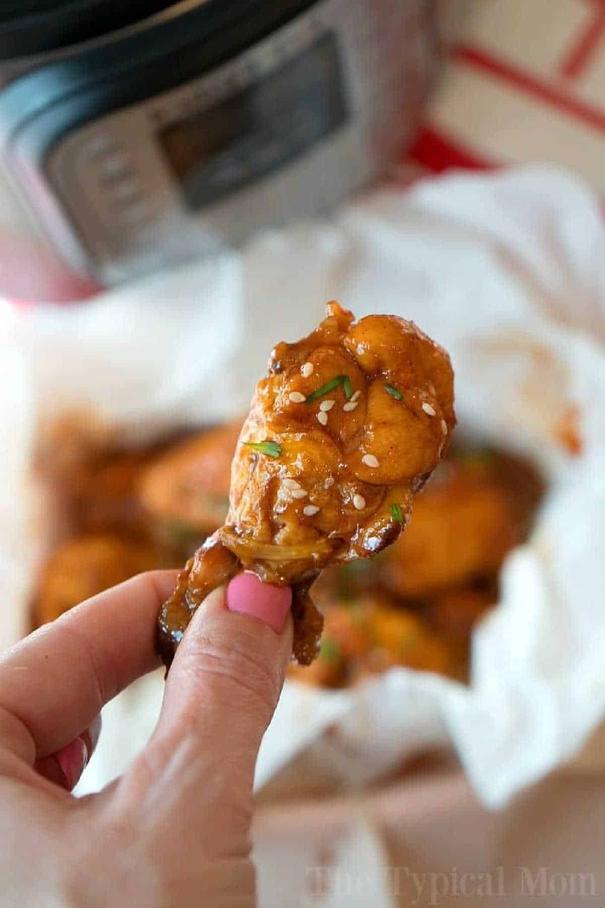 This is how you make tender and delicious pressure cooker frozen chicken wings! Makes a great appetizer or dinner in your Instant Pot using your favorite barbecue sauce. Wings are the best finger food we say and with this trick you can make them often. From freezer to dinner in less than 30 minutes! #instantpot #pressurecooker #chicken #wings #frozen #meat #time #cook A hand holds a saucy chicken wing, topped with sesame seeds and herbs—perfect inspiration for making juicy pressure cooker frozen chicken wings at home.