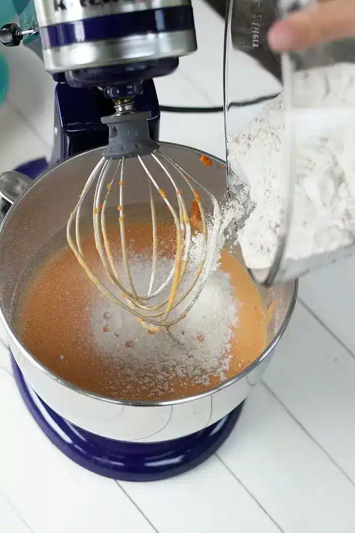 A close-up of a stand mixer with orange batter in the bowl, partially visible whisk attachment, and a glass bowl on the right pouring in white flour. 