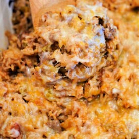 A spoon serving a cheesy baked hamburger and rice casserole with ground meat and pasta from a dish.