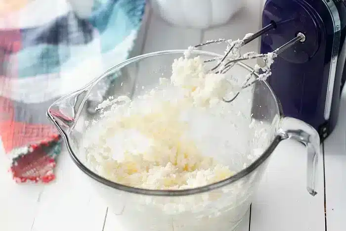 A clear glass mixing bowl containing a creamy mixture is placed on a white surface. An electric hand mixer with beaters covered in the mixture rests in the bowl, ready to whip up a 1/2 sheet cake. 