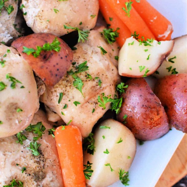 A dish of seasoned chicken thighs with baby carrots, red potatoes, and parsley garnish on a white plate, this instant pot chicken and potatoes recipe offers a quick and delectable meal thats both comforting and effortless.