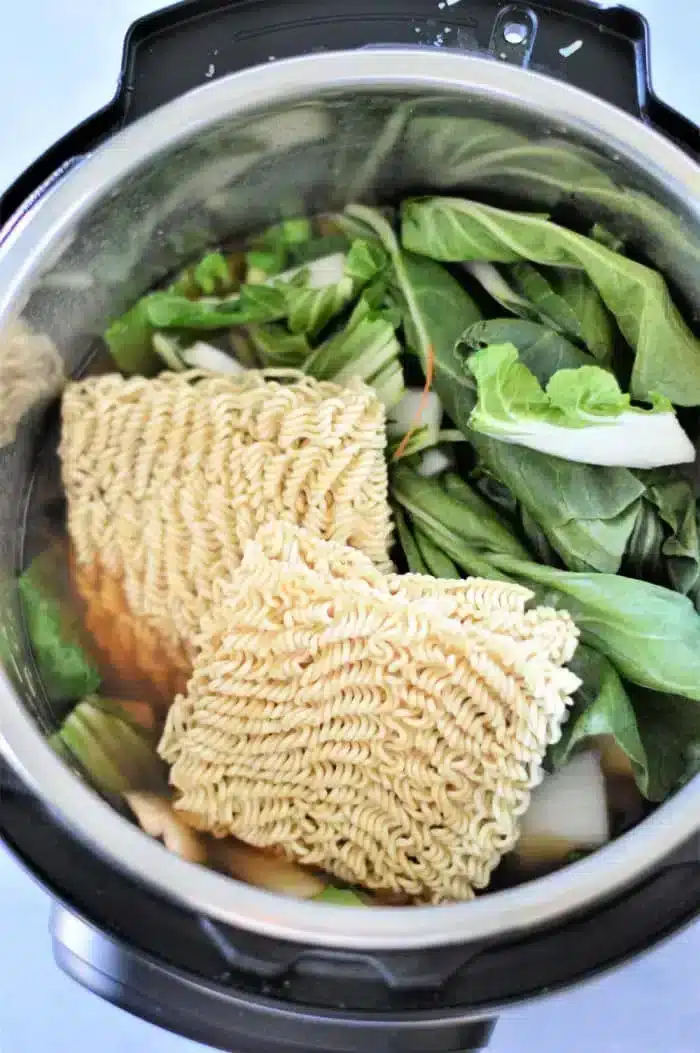 The Instant Pot is filled with vibrant ingredients for a delectable vegetable ramen. Two blocks of dry instant noodles rest atop an assortment of fresh leafy greens and sliced roots, all simmering in the savory broth.