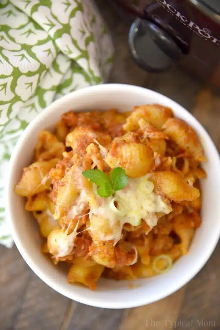 A bowl of pasta with tomato sauce, seasoned leftover taco meat, and melted cheese is garnished with a sprig of parsley. A green and white patterned cloth is visible in the background, alongside a crockpot. The bowl is placed on a wooden surface.