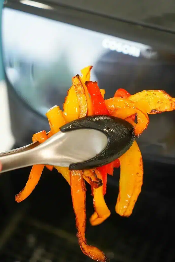 Tongs holding roasted air fryer peppers in front of an air fryer. The peppers are orange and red, with a slightly charred appearance. The air fryer is partially visible in the background.