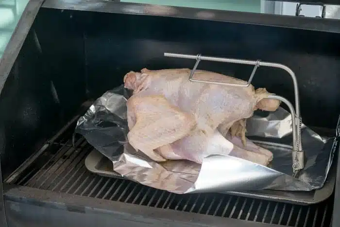 A raw turkey, generously coated with a savory rub, is placed inside a smoker on a grill with foil underneath. The lid is open, and a temperature probe is expertly inserted into the turkey.