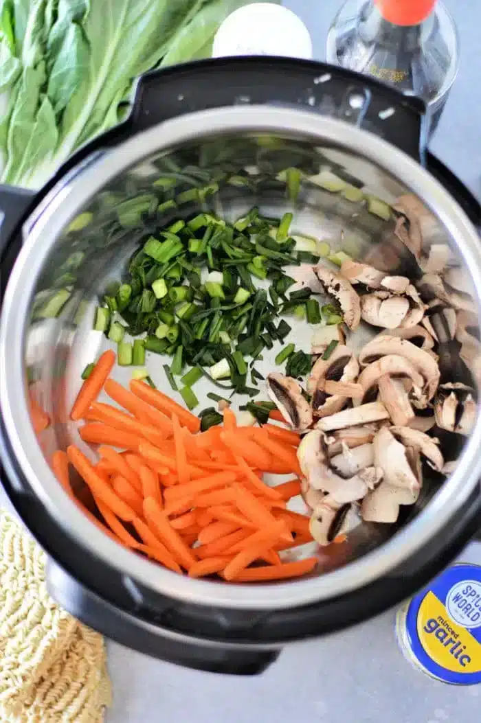 An overhead view of an Instant Pot reveals a delightful Vegetable Ramen with sliced carrots, chopped green onions, and mushrooms. Surrounding the pot are garlic, soy sauce, and leafy greens on a countertop.