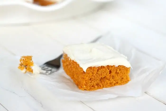 A slice of pumpkin cake with white frosting on top sits on a piece of parchment paper.