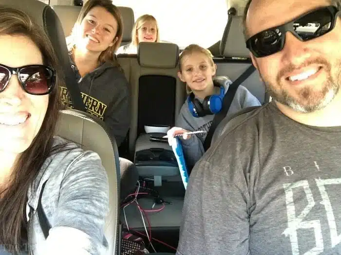 A group of five, including a man in sunglasses, a woman with brown hair, and three children enjoying road trip games for kids with headphones on, are seated inside a car. The children's smiles suggest they're having fun on their outing.