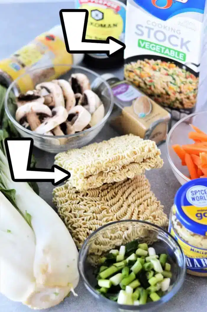 Various cooking ingredients are arranged on a surface, ready to create an Instant Pot Vegetable Ramen delight. The spread includes mushrooms in a bowl, vegetable stock, dry ramen noodles, bok choy, green onions, carrots, soy sauce, ginger, and garlic. Two arrows point toward the mushrooms and noodles.