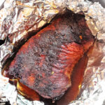 A cooked brisket is resting in a pan, wrapped partially with aluminum foil. The air fryer brisket appears well-seasoned and juicy, with a dark, caramelized exterior. There are flavorful juices collected at the bottom of the pan.