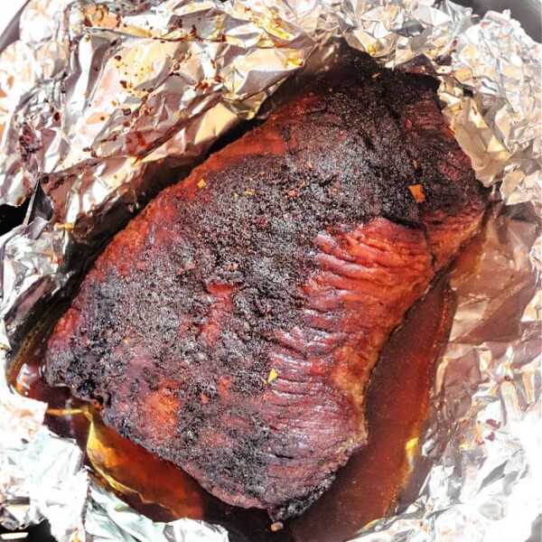 A cooked brisket is resting in a pan, wrapped partially with aluminum foil. The air fryer brisket appears well-seasoned and juicy, with a dark, caramelized exterior. There are flavorful juices collected at the bottom of the pan.