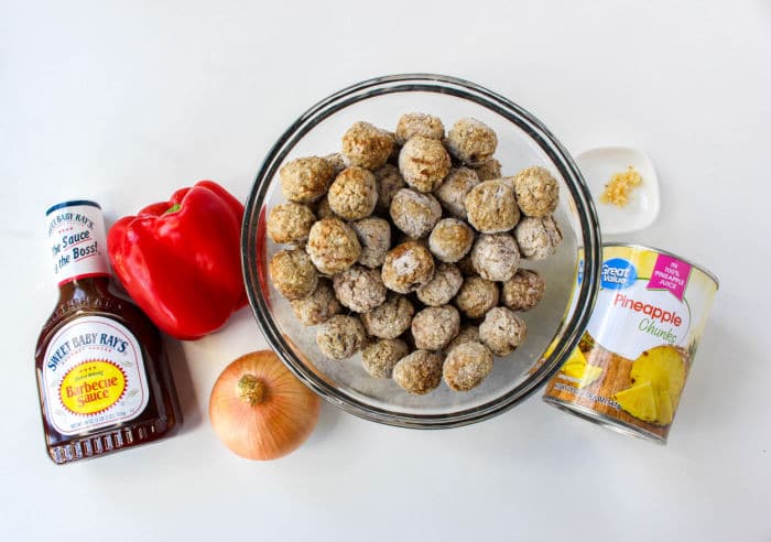 A bowl of frozen meatballs, perfect for Slow Cooker BBQ Meatballs, is surrounded by a bottle of barbecue sauce, a red bell pepper, an onion, a can of pineapple chunks, and minced garlic on a white surface.