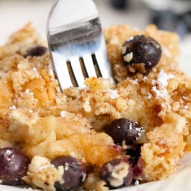 Close-up of a fork piercing a slice of blueberry bread pudding, its crumb-topped layers offering a delectable treat on a white plate.