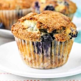 A close-up of a jumbo blueberry muffin on a white plate. The muffin has a golden-brown top with visible blueberry pieces and a slightly crumbly texture. Another jumbo muffin is partially visible in the background. The plate is on a white surface with an orange-striped cloth.