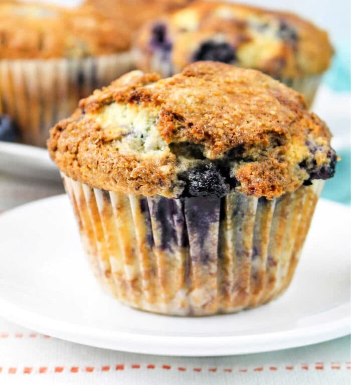 A close-up of a jumbo blueberry muffin on a white plate. The muffin has a golden-brown top with visible blueberry pieces and a slightly crumbly texture. Another jumbo muffin is partially visible in the background. The plate is on a white surface with an orange-striped cloth.