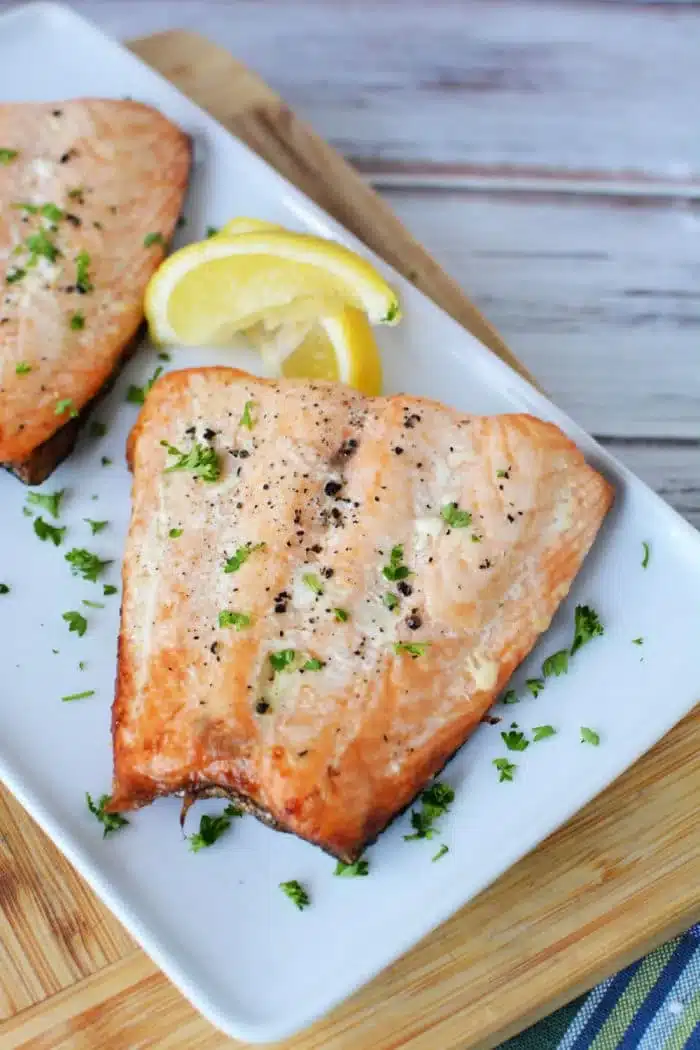 A delectable grilled salmon fillet rests on a rectangular white plate, garnished with chopped parsley and black pepper. Two lemon wedges accompany this delight. The plate, showcasing perfectly thawed frozen salmon grill markings, sits on a wooden cutting board with a striped cloth beneath.