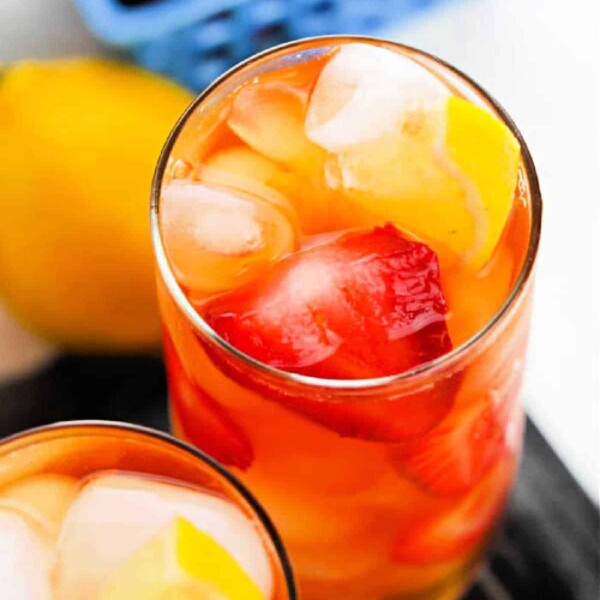 Two glasses of iced hibiscus sweet tea with strawberries and lemon slices, accompanied by a basket of fresh strawberries in the background.