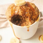 Banana Bread in a Mug: A mouthwatering banana mug cake with a bite taken, topped with fresh banana slices and drizzled syrup, all on a pristine white surface.