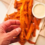 A hand holds a perfectly crispy fry above a plate brimming with frozen sweet potato fries, fresh from the air fryer, next to a tempting bowl of dipping sauce.
