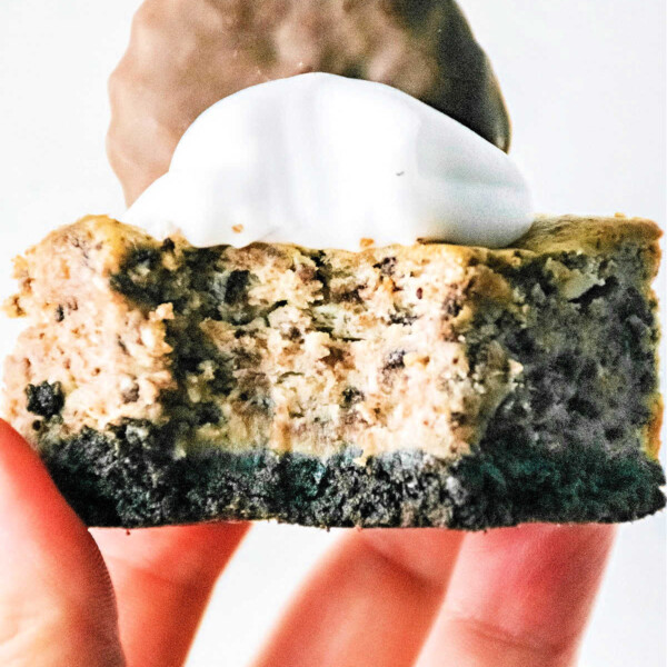 A hand holding a delectable slice of Oreo cheesecake, reminiscent of Philadelphia Cheesecake Bars, topped with whipped cream and a chocolate biscuit.