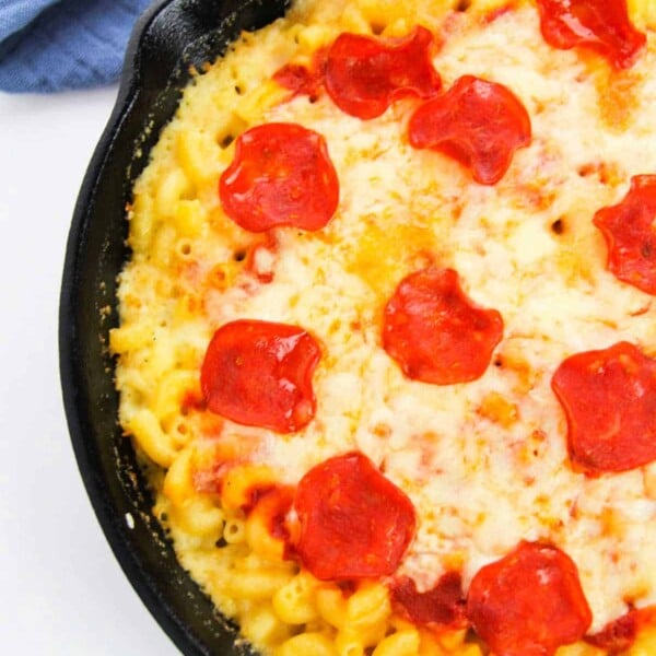 A skillet of Pizza Mac and Cheese features cheesy macaroni topped with pepperoni slices, resting next to a blue cloth.