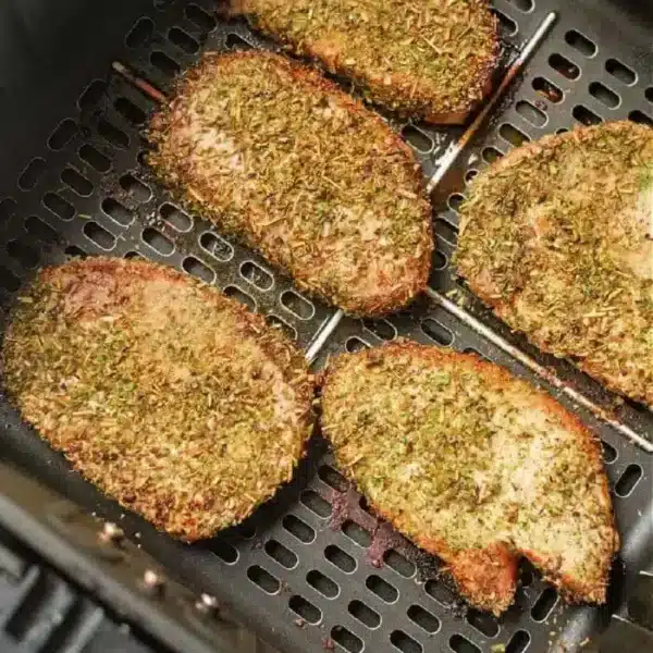 Five seasoned pork steaks are evenly spaced in an air fryer basket. They are covered in a mixture of herbs and spices, giving them a crispy, golden-brown appearance.