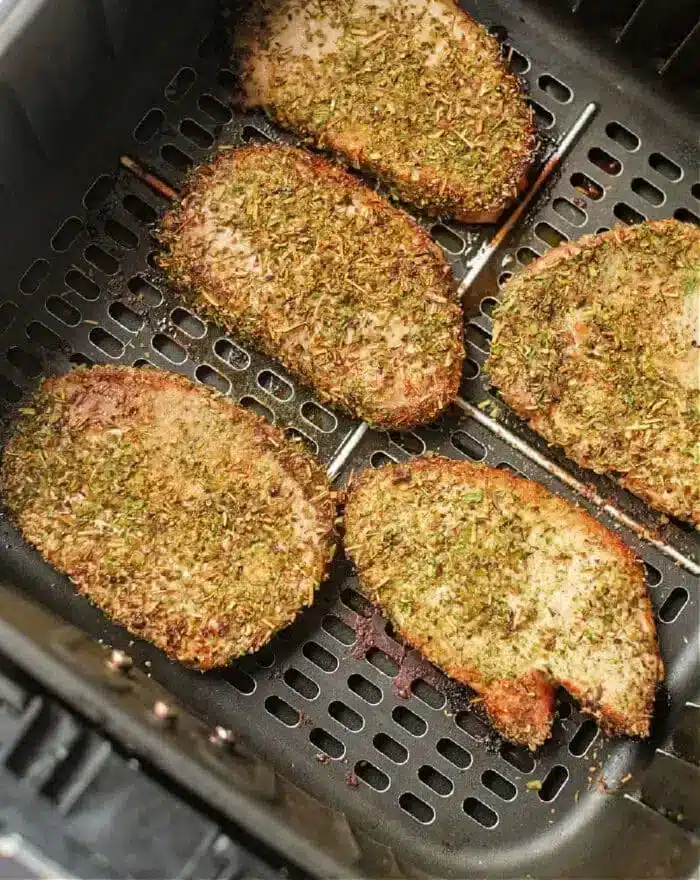 Five seasoned pork steaks are evenly spaced in an air fryer basket. They are covered in a mixture of herbs and spices, giving them a crispy, golden-brown appearance.