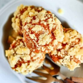 A plate contains three crumb-topped mini pumpkin muffins, showcasing a golden-brown streusel layer. A fork with a wooden handle is partially visible next to the muffins, suggesting readiness for eating. The background is blurred, emphasizing the food as the main focus.