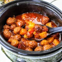 Slow cooker BBQ meatballs and pineapple chunks swim in a tangy red sauce, with a ladle resting inside. Pineapple peeks from the background, promising that sweet and savory magic awaits.