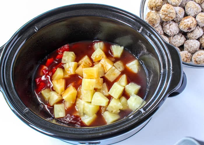 A slow cooker brimming with a tangy red sauce spiced with diced pineapples and red peppers. Beside it, on the pristine white surface, rests a bowl of uncooked BBQ meatballs, ready to transform into savory delights.