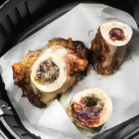 Three pieces of roasted air fryer bone marrow sit perfectly on a sheet of parchment paper inside the basket, their surfaces golden and slightly charred.