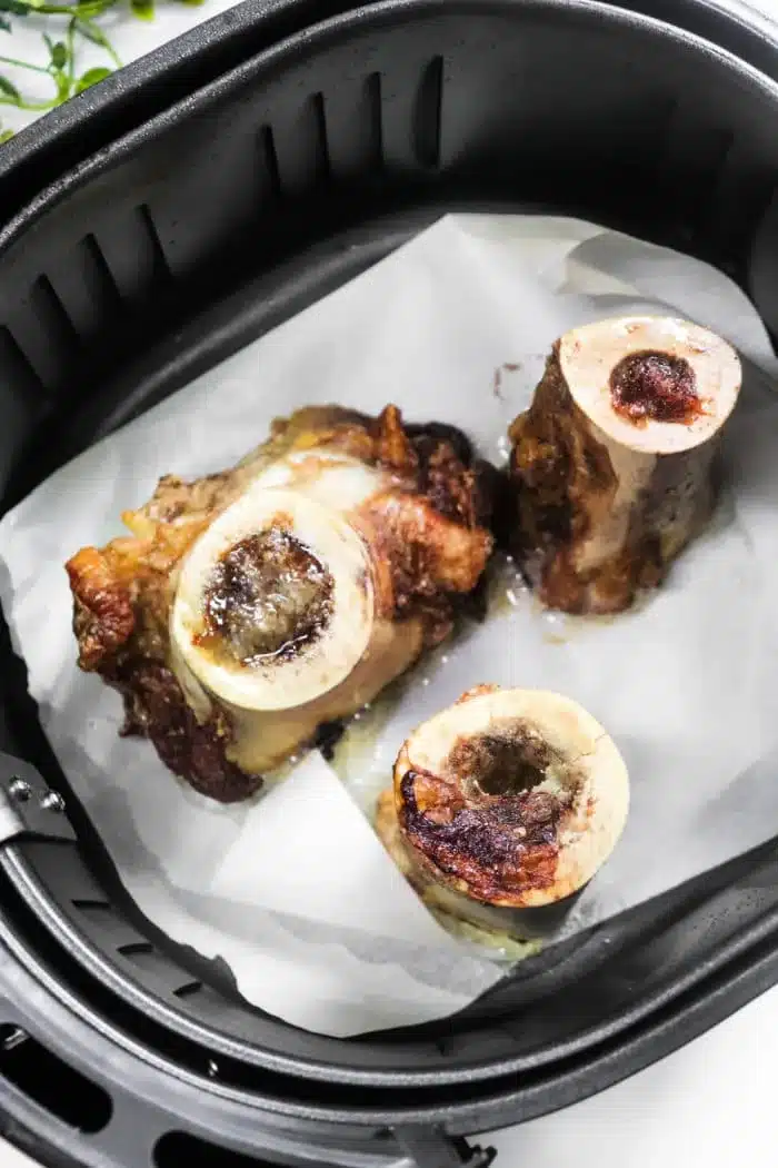 Three pieces of roasted air fryer bone marrow sit perfectly on a sheet of parchment paper inside the basket, their surfaces golden and slightly charred.