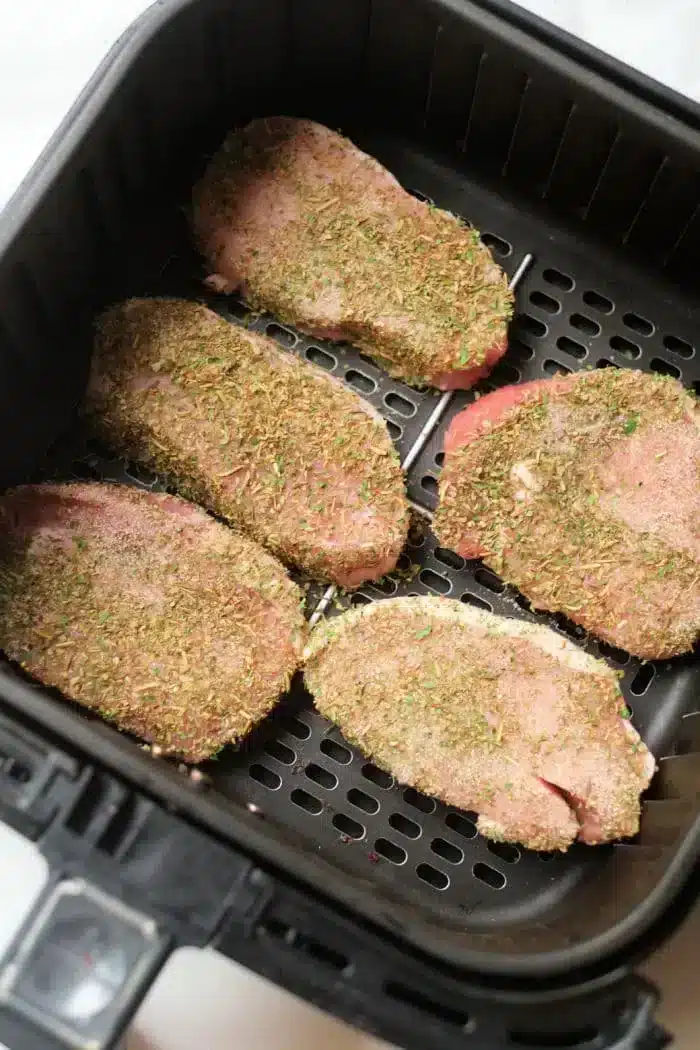 Five uncooked, seasoned pork steaks are placed in an air fryer basket, ready for cooking. The meat is covered with a mixture of herbs and spices.