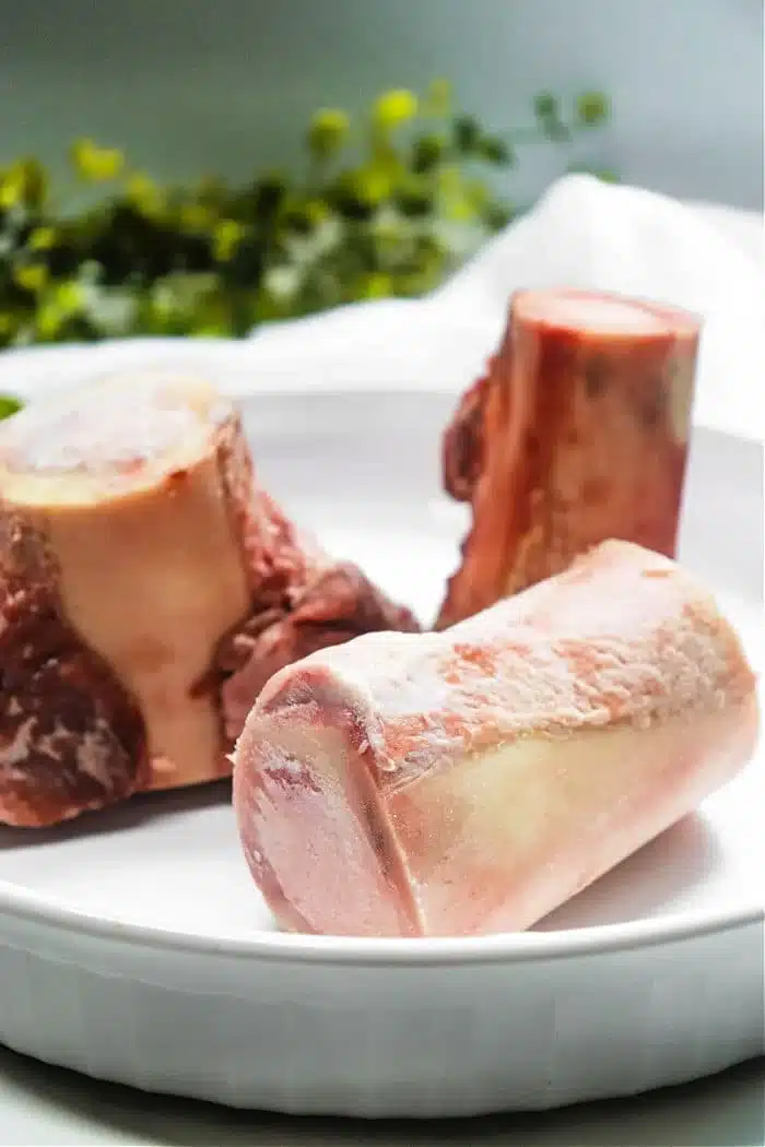 Three raw beef bones, perfect for air fryer bone marrow, are placed in a white dish. The bones have a light pink hue with some white marbling and fat visible. They are situated in a well-lit setting with green foliage blurred in the background.