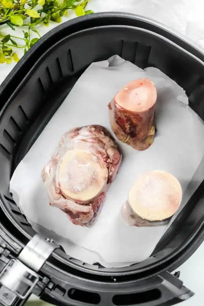 Juicy beef marrow bones rest on parchment paper inside an air fryer basket, ready to become a gourmet delight. A few green leaves peek through in the background, hinting at a fresh twist to this classic air fryer bone marrow delicacy.