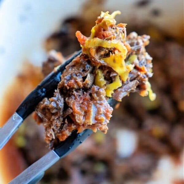 A fork holds a portion of shredded beef mixed with melted cheese and chopped vegetables. The background is blurred, showing more of the same mixture in a dish.