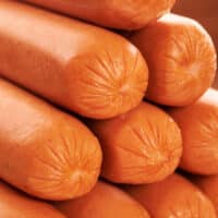 A close-up image reveals a stack of smooth, orange-colored hot dog sausages, their ends sealed and slightly pinched. Tightly packed to showcase their glossy texture and cylindrical shape, these sausages prompt the question: how long to boil hot dogs for perfect tenderness?