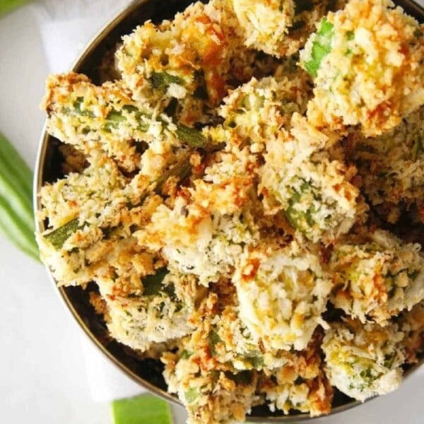 A bowl of air fryer okra, golden and crispy, sits on a white surface, with a few fresh okra pods nearby.