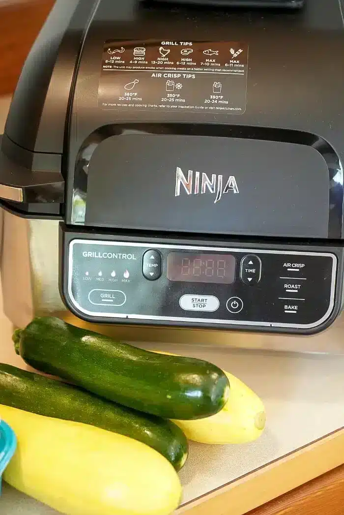 A Ninja air fryer sits on a kitchen counter, ready to effortlessly prepare air fryer vegetables with two zucchinis and a yellow squash in front of it. The control panel boasts various options on its digital screen, while grill and air crisp instructions are neatly labeled on the lid.