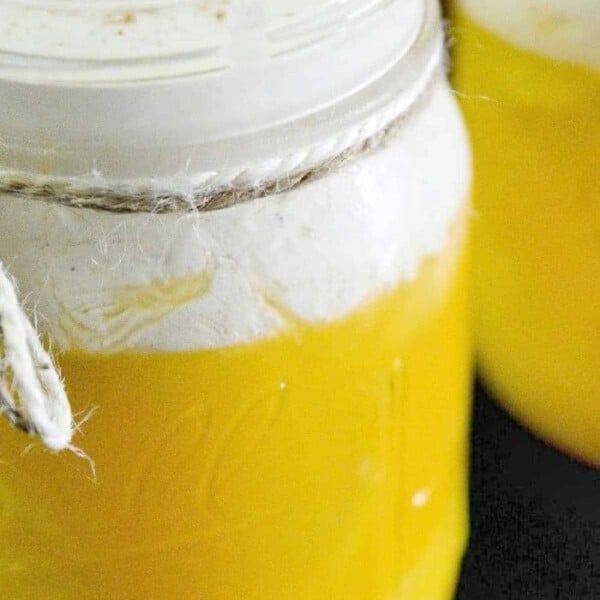 Close-up of a mason jar filled with what resembles an orange creamsicle drink, its vibrant yellow liquid topped with thick white foam and a sprinkle of cinnamon. The jar is elegantly tied with a piece of twine around its neck.